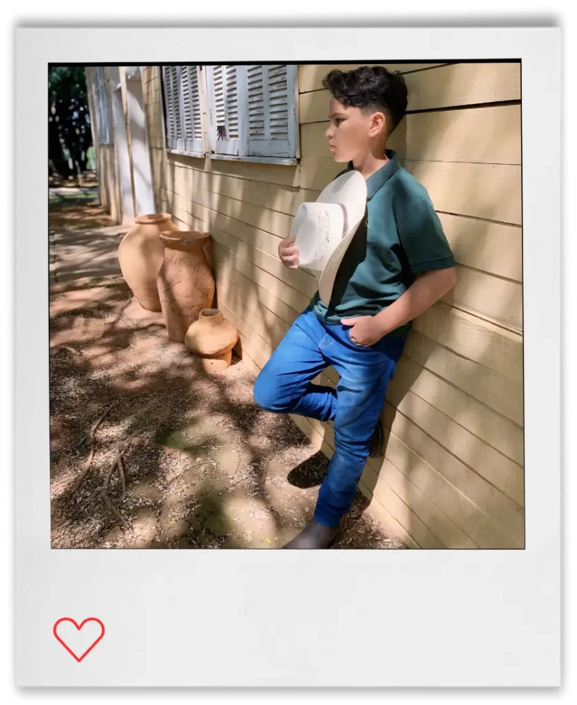 O jovem artista João Pedro Violeiro em foto estilo sertanejo, encostado em uma casa de fazenda e segurando um chapéu.